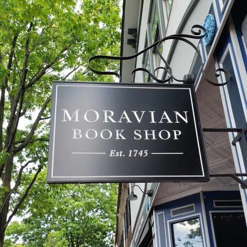 Sign for Moravian Book Shop, established in 1745, hanging from a building with green trees in the background.