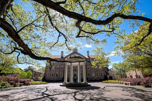 A gazebo surrounded by blooming flowers and trees, with a historic building in the background under a clear blue sky.