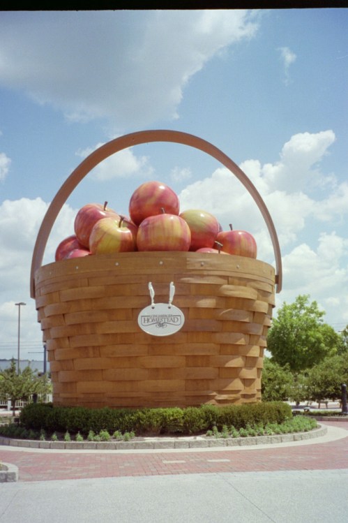 A large wooden basket overflowing with oversized apples, set against a blue sky with fluffy clouds.