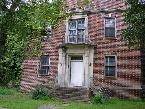 Abandoned brick building with a white door, stairs, and overgrown greenery surrounding it.