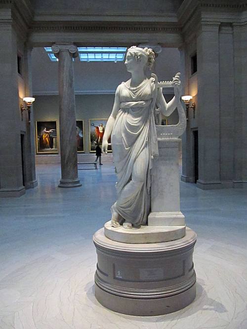 A marble statue of a woman holding a lyre, set in a spacious gallery with columns and paintings in the background.