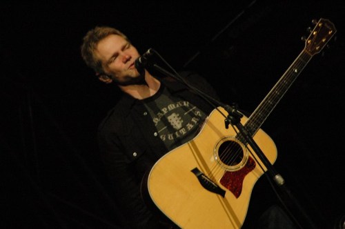 A musician smiles while singing into a microphone, holding an acoustic guitar on stage.