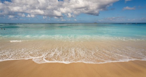 A serene beach scene with gentle waves lapping at the sandy shore under a partly cloudy blue sky.