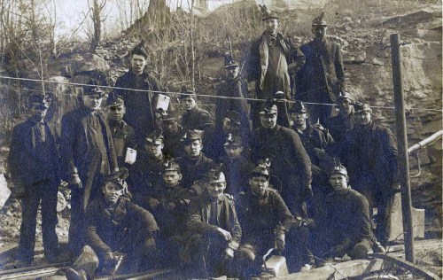 A historical black-and-white photo of a group of miners posing together outdoors, wearing helmets and work clothes.