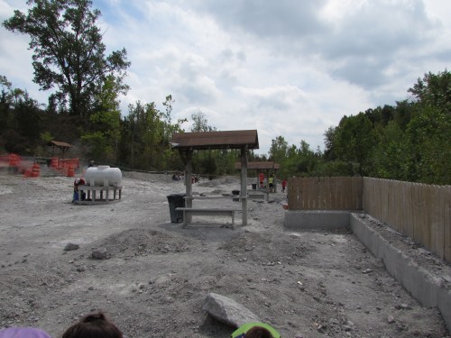 A gravel site with wooden shelters, water tanks, and trees in the background under a cloudy sky.