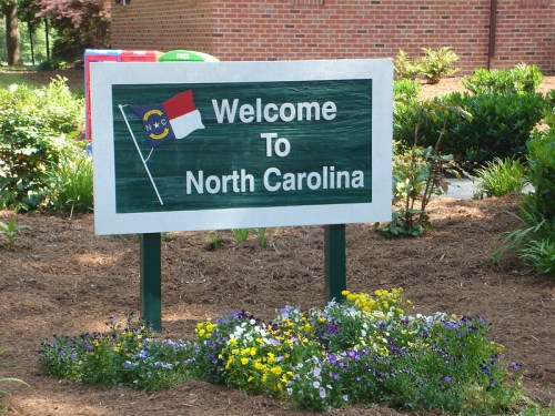 Welcome sign for North Carolina, featuring the state flag and surrounded by colorful flowers and greenery.