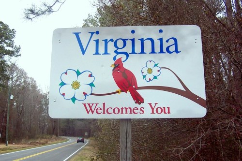 Sign reading "Virginia Welcomes You" featuring a cardinal and dogwood flowers, set against a wooded background.
