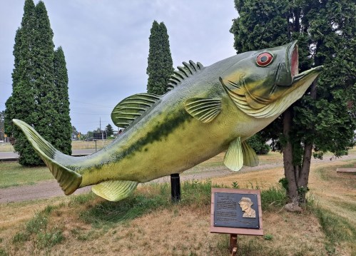 A large, colorful statue of a fish stands beside a plaque, surrounded by trees and grass under a cloudy sky.