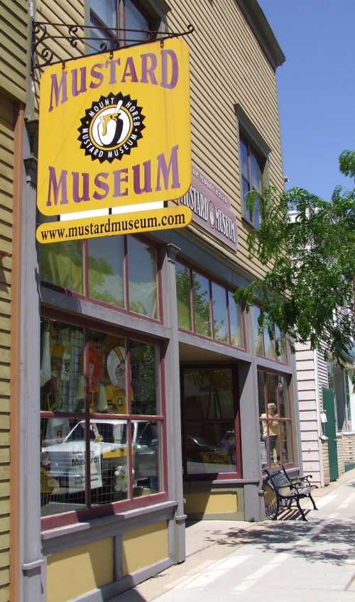 Exterior of the Mustard Museum, featuring a large sign and colorful storefront with windows and a bench outside.