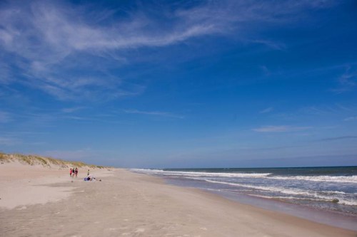 A sandy beach stretches under a blue sky, with gentle waves and a few people enjoying the shoreline.