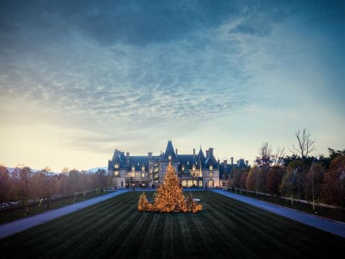 A grand mansion at dusk, surrounded by a manicured lawn and trees, with warm lights glowing from the windows.