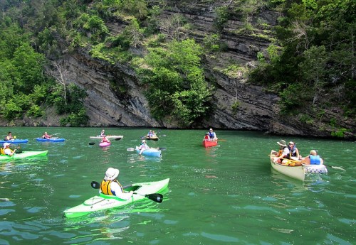 A group of kayakers in colorful kayaks paddling on a calm green lake surrounded by rocky cliffs and lush greenery.
