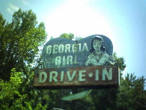 Vintage sign for "Georgia Girl Drive-In" featuring a smiling girl illustration against a blue sky.