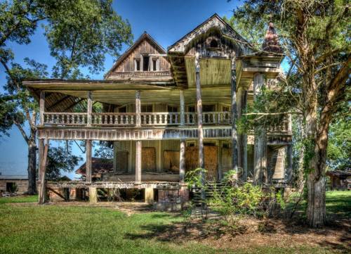 An abandoned, weathered house with a large porch, surrounded by trees and overgrown grass under a clear blue sky.