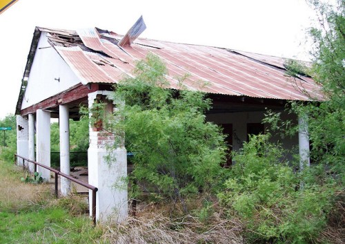 Abandoned building with a rusted roof, overgrown vegetation, and crumbling structure in a rural setting.