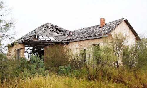 An abandoned, dilapidated house with a collapsed roof, surrounded by overgrown grass and shrubs.