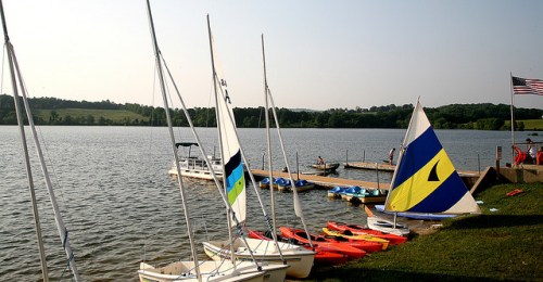 Sailboats and kayaks lined up by a calm lake, with a dock and an American flag in the background.