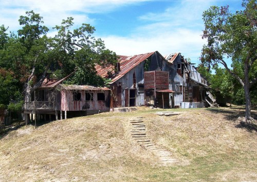 An old, weathered building with a rusty roof, surrounded by trees and grass, sits on a hill under a blue sky.