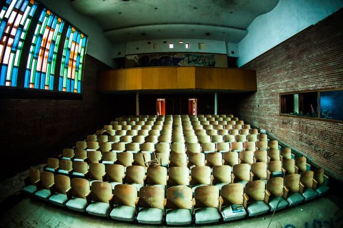 Abandoned auditorium with rows of empty, worn seats and a colorful stained glass window.