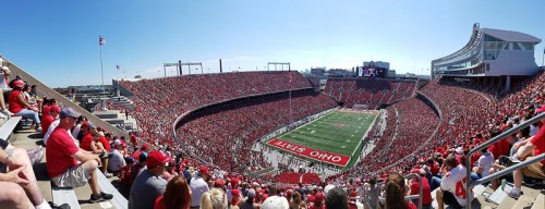 A panoramic view of a packed stadium filled with fans in red, watching a football game under a clear blue sky.