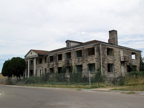 Abandoned stone building with broken windows, overgrown vegetation, and a chimney, set against a cloudy sky.