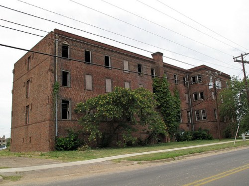 Abandoned brick building with broken windows, overgrown vegetation, and power lines in a rural setting.