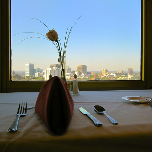 Table setting with a pink napkin and utensils, overlooking a city skyline and blue sky through a large window.
