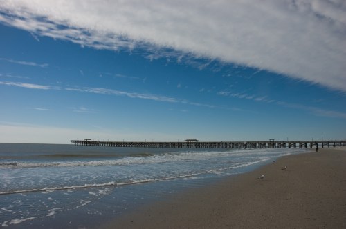 A serene beach scene featuring a long pier extending into calm waters under a blue sky with wispy clouds.