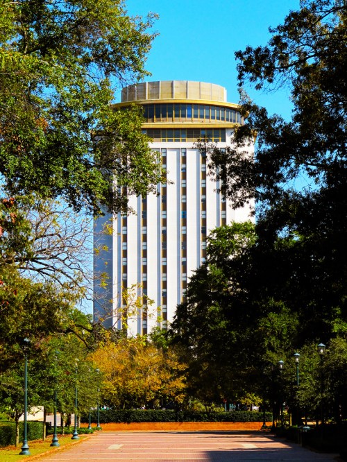 A tall building with a circular top, surrounded by trees and a clear blue sky, viewed from a pathway.