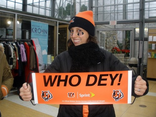A woman in a Bengals cap and scarf holds a sign that says "WHO DEY!" in a greenhouse setting.