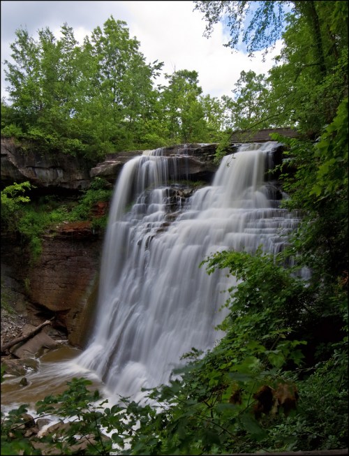 A cascading waterfall surrounded by lush green trees and rocky cliffs under a cloudy sky.