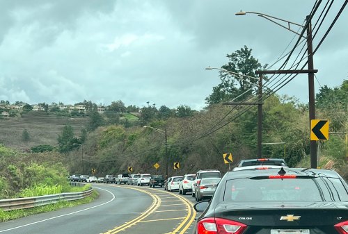 Traffic jam on a winding road surrounded by greenery and power lines under a cloudy sky.