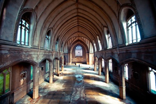 A spacious, abandoned church interior with high arches, large windows, and sunlight reflecting on the wet floor.