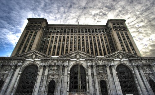 A large, abandoned building with ornate architecture under a dramatic sky, featuring tall windows and intricate details.