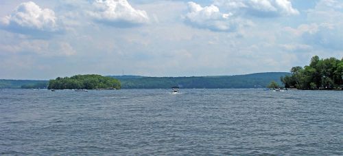 A serene lake scene with boats on the water, surrounded by green trees and distant hills under a cloudy sky.