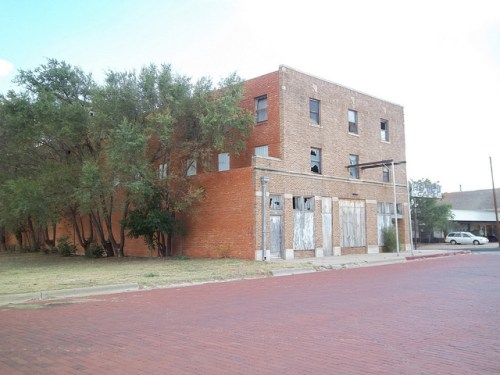 Abandoned brick building with boarded windows, surrounded by trees, on a quiet street with red brick pavement.
