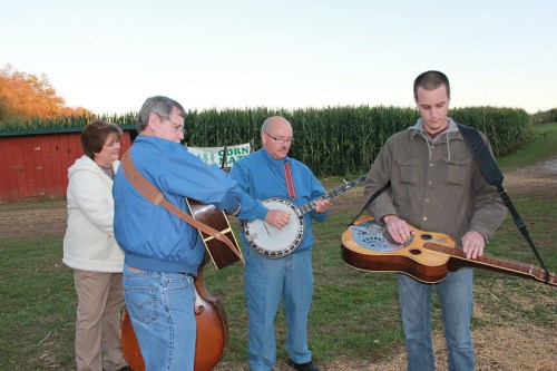 Four musicians play instruments outdoors near a cornfield, with a red barn in the background.