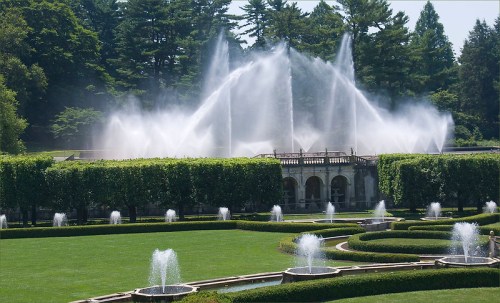 A grand fountain sprays water in a lush garden, surrounded by neatly trimmed hedges and vibrant greenery.