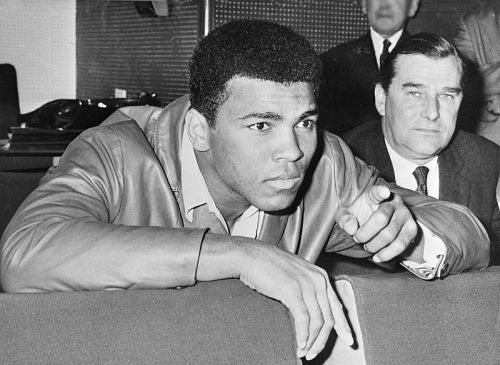 A young man in a leather jacket leans forward, pointing, with a serious expression; a man in a suit sits behind him.