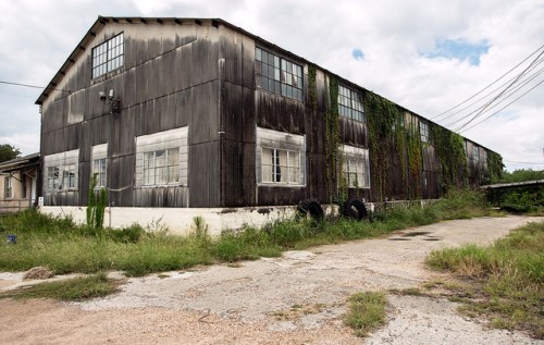 Abandoned industrial building with weathered wood, overgrown vegetation, and a gravel path in front.