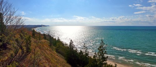 A panoramic view of a shimmering lake with gentle waves, framed by trees and a clear blue sky.
