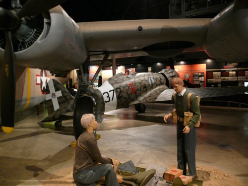 Two life-sized figures in military attire interact in front of a vintage aircraft in a museum setting.