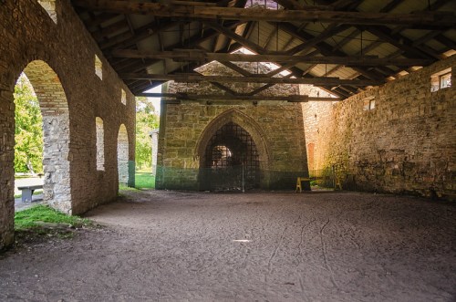 Interior of a stone building with arched windows and a wooden roof, featuring a dirt floor and a large archway.