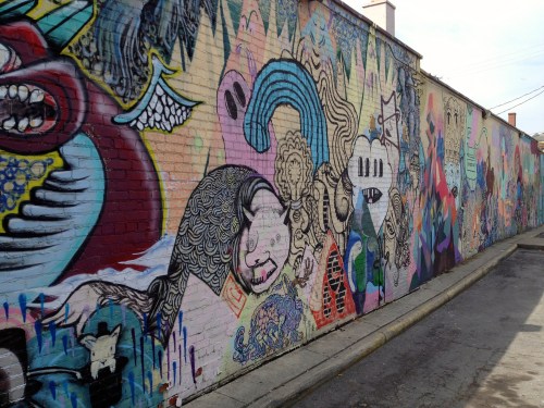 Colorful street mural featuring abstract characters and intricate designs on a brick wall in an alleyway.