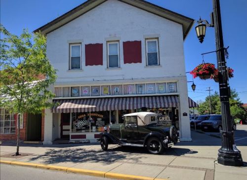 A vintage car parked in front of a quaint building with a striped awning and flower baskets on a sunny day.