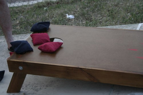 A wooden cornhole board with four bean bags resting on it, set on grass with a fence in the background.