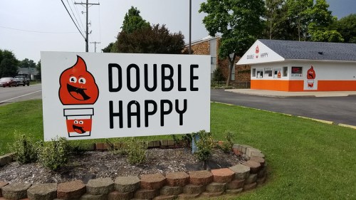 A sign for "Double Happy" featuring a smiling flame logo, with a small building in the background. Green grass surrounds the area.