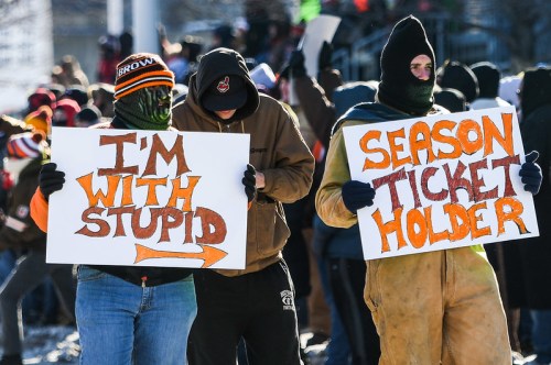 Two individuals in winter clothing hold signs at a protest. One sign reads "I'M WITH STUPID" and the other "SEASON TICKET HOLDER."