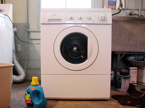A white front-loading washing machine in a laundry room, with a bottle of laundry detergent in front.