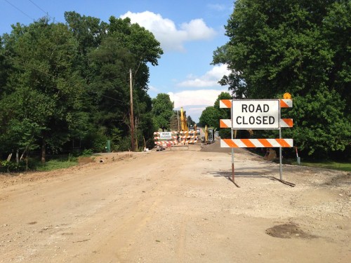 A dirt road is closed with a "Road Closed" sign and construction barriers in a wooded area under a blue sky.